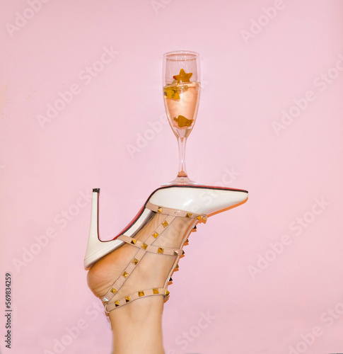 Creative photo of a woman's foot in white shoes, with a glass of champagne on the shoe, on a pink background. The concept of Valentine's Day.
