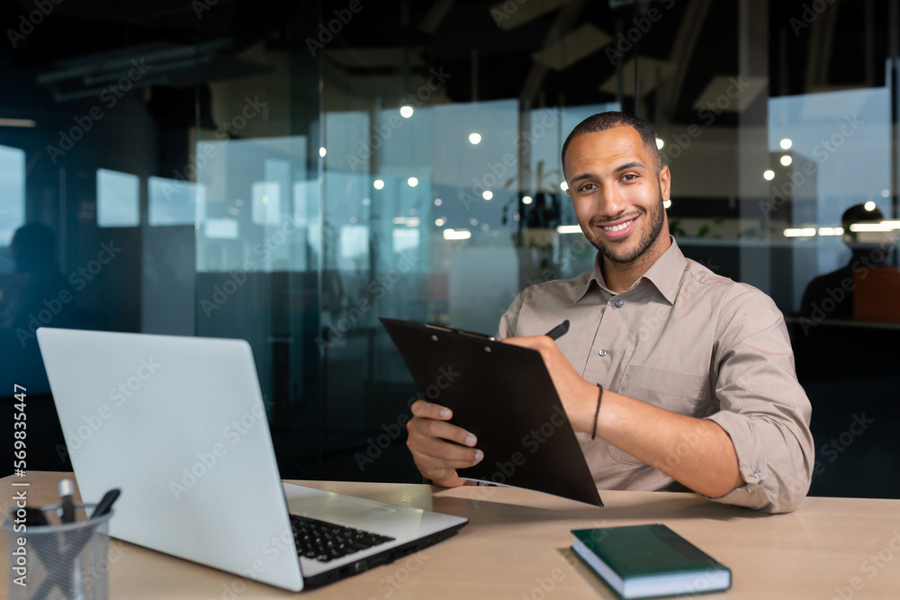 Portrait of smiling and satisfied businessman with achievement results, african american paper work smiling and looking at camera, young financier writing document inside office using laptop and pen. Portrait of smiling and satisfied businessman with achievement results, african american paper work smiling and looking at camera, young financier writing document inside office using laptop and pen.