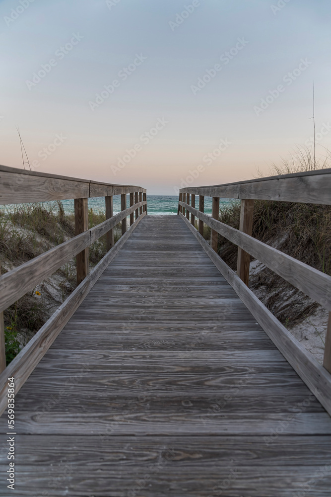 Vertical shot view of a wooden pathway with railings in between grassy sand dunes in Destin, Florida. Pathway heading to the beack with blue ocean under the horizon skyline.