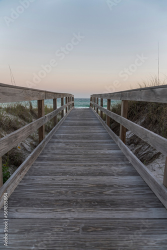 Vertical shot view of a wooden pathway with railings in between grassy sand dunes in Destin  Florida. Pathway heading to the beack with blue ocean under the horizon skyline.
