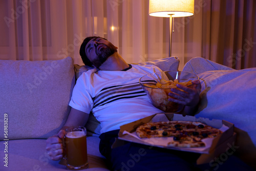 Man with chips, pizza and glass of beer sleeping on sofa at night. Bad habit