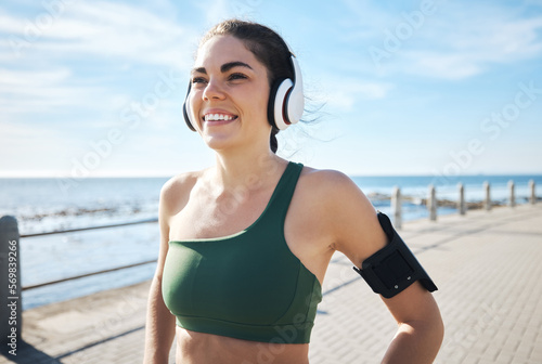 Happy, fitness and woman with music at beach for running, exercise and cardio on blue sky background. Radio, podcast and workout run by girl at ocean for training, smile or relax in audio motivation