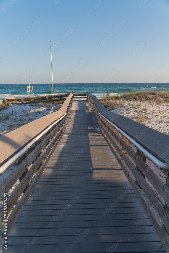 Wooden pathway along with the grassy sand dunes heading to the beach at Destin, Florida. Footbridge with railings and views of blue ocean and skyline background.