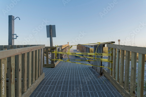 Closed part of a boardwalk with yellow caution tape tied on the wooden railings in Destin, Florida. Screen walkway of a boardwalk with shower station on the left side. © Jason