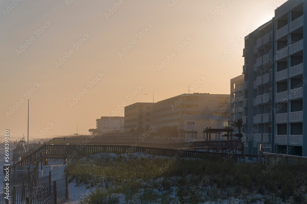 Sunset behind the row of the hotel buildings with grassy sand dunes at the front in Destin, Florida. There are wooden fences and sand dunes with footbridges connecting the beach and buildings.