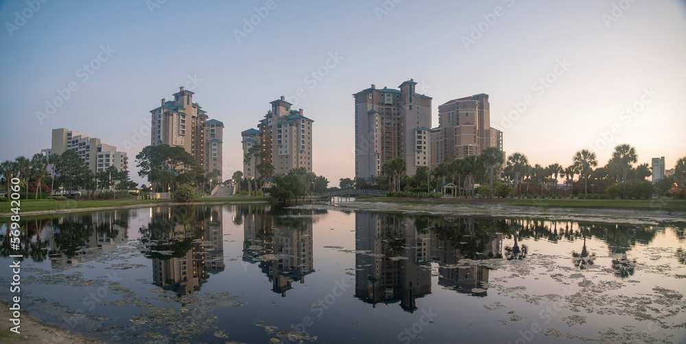 Fototapeta premium Panorama of decorative lake in a golf course with reflections of hotel buildings at Destin, Florida. Cityscape of hotels view from a golf course against the dusk sky.