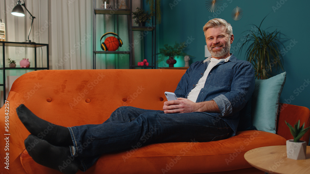Fototapeta premium Mature senior bearded man lying on sofa uses mobile phone smiles at night evening home. Mature guy texting share messages content on smartphone social media applications online watching relax movie