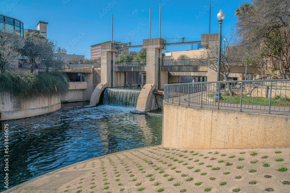 View of an open flood gates at San Antonio River from a boat ramp with grass pavement at River Walk. There is a view of a pathway with railings on the right and bridge over the flood gates.