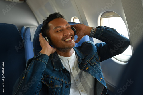 Happy asian man in wireless headphones listening to music and sitting comfortable seat in airplane cabin