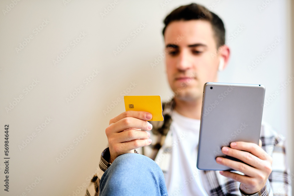 Young man shopping and paying online on digital tablet with credit card while relaxing at home listening music with earphones. Copy space. Young man shopping and paying online on digital tablet with credit card while relaxing at home listening music with earphones. Copy space.