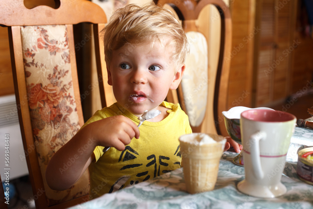 A blond boy sits at the table and eats ice cream himself with a spoon A blond boy sits at the table and eats ice cream himself with a spoon
