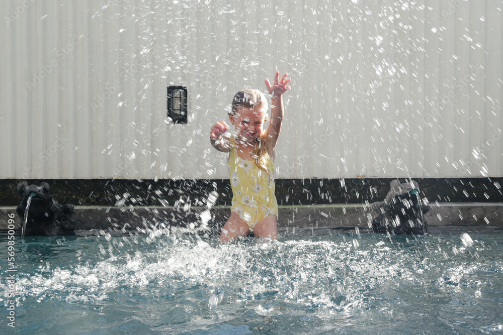 Fototapeta premium Portrait of adorable 5 years old girl sitting at the edge of swimming pool and splashing water with hands