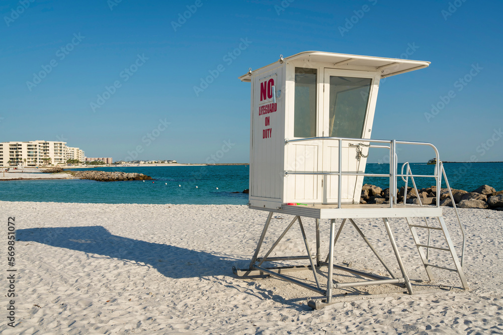 Small white lifeguard tower on the white sand shore with views of buildings and sky at Destin, FL. There are views of modern multi-storey buildings on the left across the water from a shore.