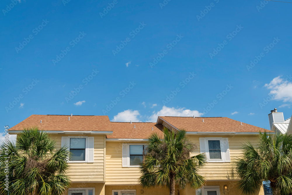 Residential building with light yellow wood lap siding and brown orange roof at Destin, Florida. Apartment house with three front doors and palm trees at the front.