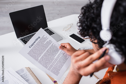 Devices near blurred indian freelancer in headphones working with paper with copywriting lettering at home.