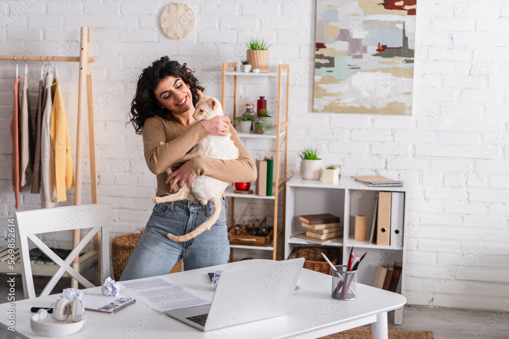 Smiling copywriter holding oriental cat near laptop and papers at home.