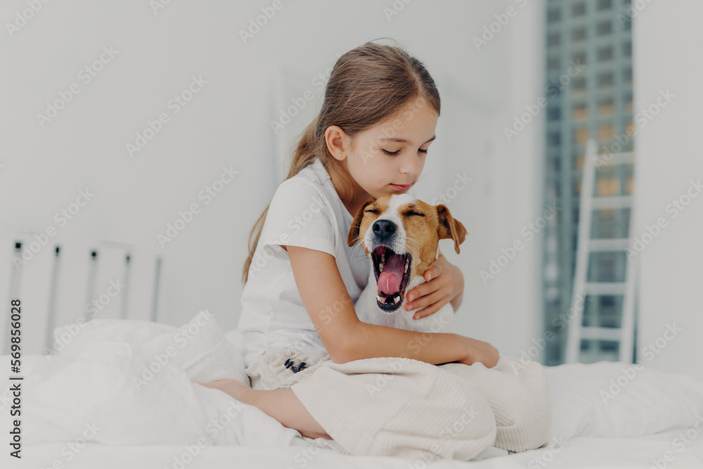 Fair haired small girl with pony tail wears casual pyjamas, embraces dog who yawns, pose together on comfortable bed, expresses love and care to domestic animal, spends morning weekend at home Fair haired small girl with pony tail wears casual pyjamas, embraces dog who yawns, pose together on comfortable bed, expresses love and care to domestic animal, spends morning weekend at home
