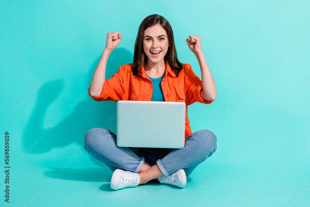 Full length photo of cute lucky lady wear orange shirt rising fists typing modern gadget isolated teal color background Full length photo of cute lucky lady wear orange shirt rising fists typing modern gadget isolated teal color background