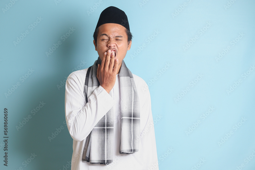 Portrait of attractive Asian muslim man in white shirt yawning with hand covering his mouth. Isolated image on blue background Portrait of attractive Asian muslim man in white shirt yawning with hand covering his mouth. Isolated image on blue background