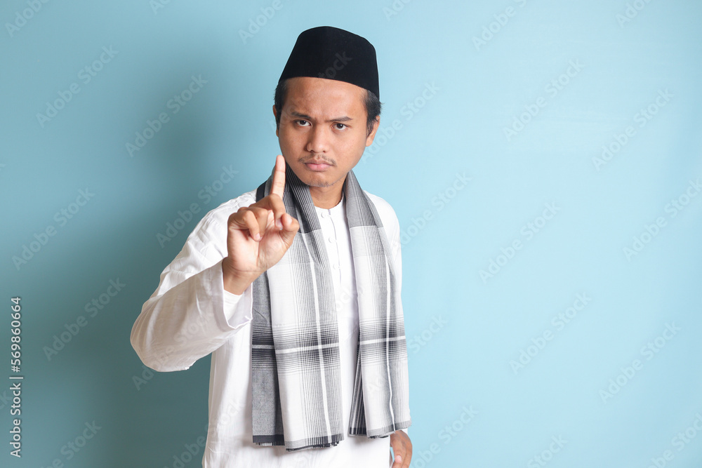 Portrait of young Asian muslim man pointing up his forefinger and reminding something to do. Advertising concept. Isolated image on blue background Portrait of young Asian muslim man pointing up his forefinger and reminding something to do. Advertising concept. Isolated image on blue background