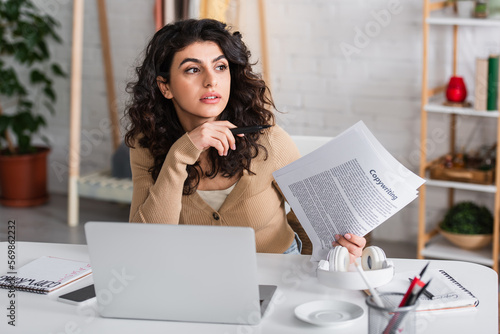 Brunette copywriter holding papers near gadgets and headphones in living room.