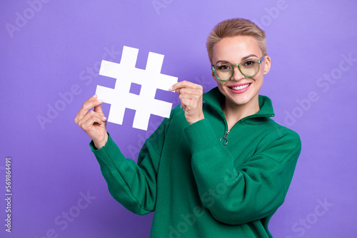Portrait photo of pretty girl wearing green fleece zipper jacket hold paper white hashtag popularity pin isolated on purple color background
