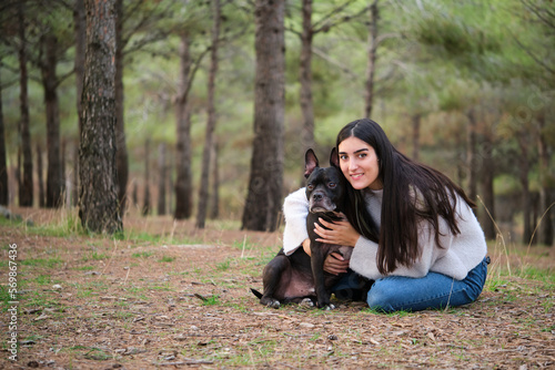 Young caucasian woman hugging her american staffordshire and french bulldog mixed breed dog and looking at camera at a pine forest.