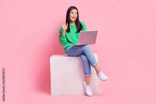 Full size portrait of nice cheerful girl sit podium arm waving communicate video call netbook isolated on pink color background