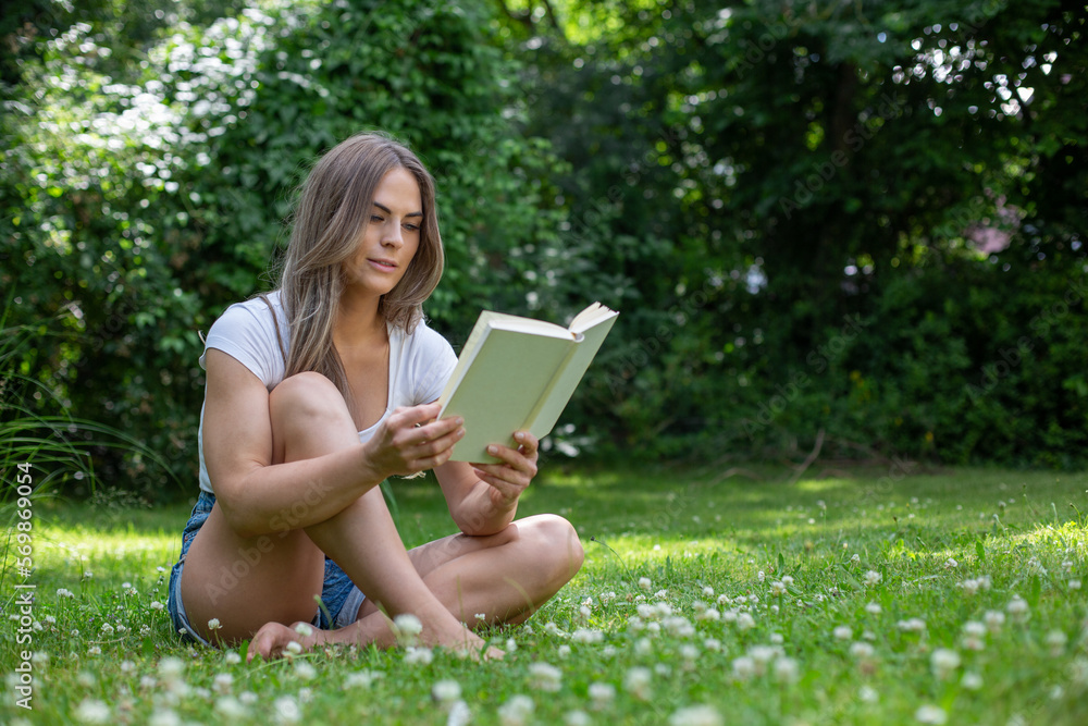 Young blond woman sits cross-legged in the garden between many daisies, reads a book and smiles Young blond woman sits cross-legged in the garden between many daisies, reads a book and smiles