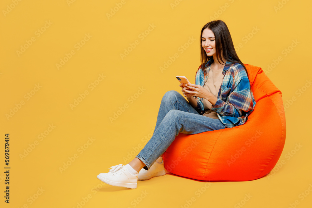 Full body young happy fun smiling woman wear blue shirt beige t-shirt sit in bag chair hold in hand use mobile cell phone isolated on plain yellow background studio portrait. People lifestyle concept. Full body young happy fun smiling woman wear blue shirt beige t-shirt sit in bag chair hold in hand use mobile cell phone isolated on plain yellow background studio portrait. People lifestyle concept.