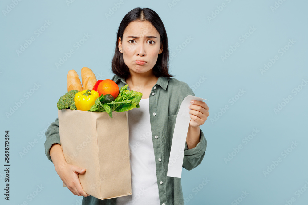 Young shocked sad woman wearing casual clothes hold brown paper bag with food products look at check isolated on plain blue cyan background studio portrait. Delivery service from shop or restaurant. Young shocked sad woman wearing casual clothes hold brown paper bag with food products look at check isolated on plain blue cyan background studio portrait. Delivery service from shop or restaurant.