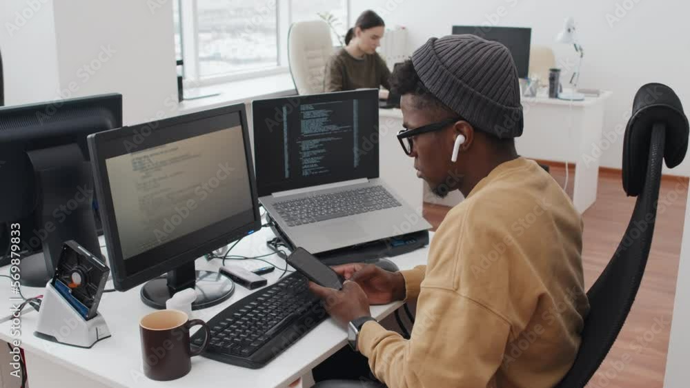 Serious young Black cybersecurity specialist looking at computer screen and texting message to colleague using smartphone Serious young Black cybersecurity specialist looking at computer screen and texting message to colleague using smartphone