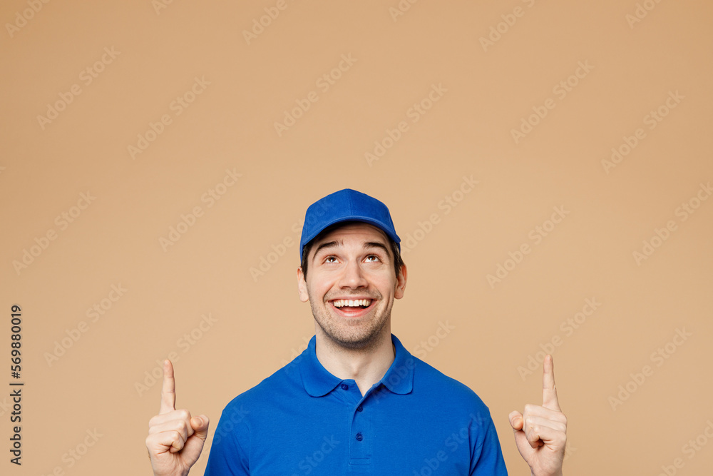 Fun delivery guy employee man wearing blue cap t-shirt uniform workwear work as dealer courier point index finger overhead indicate on area isolated on plain light beige background. Service concept. Fun delivery guy employee man wearing blue cap t-shirt uniform workwear work as dealer courier point index finger overhead indicate on area isolated on plain light beige background. Service concept.