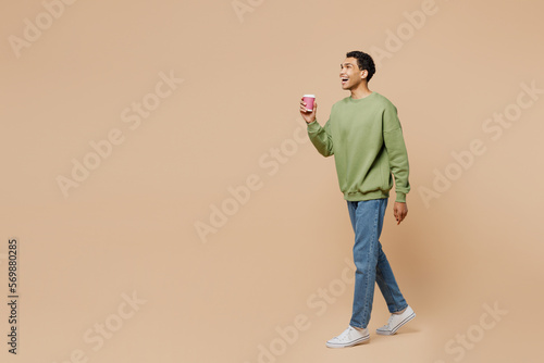 Full body young man of African American ethnicity wear green sweatshirt hold takeaway delivery craft paper brown cup coffee to go isolated on plain pastel beige background. People lifestyle concept.