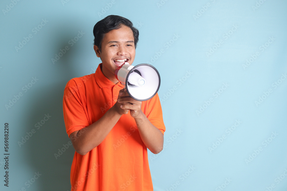 Portrait of attractive Asian man in orange shirt speaking louder using megaphone, promoting product. Advertising concept. Isolated image on blue background Portrait of attractive Asian man in orange shirt speaking louder using megaphone, promoting product. Advertising concept. Isolated image on blue background