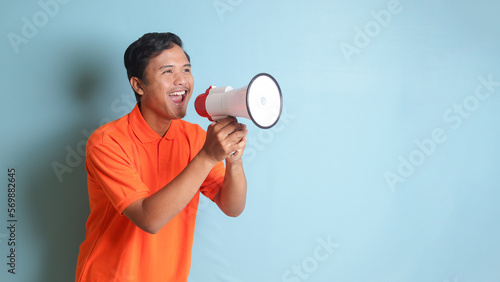 Bilde på lerret Portrait of attractive Asian man in orange shirt speaking louder using megaphone, promoting product