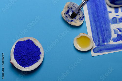 Pigmento azul y aceite mezclados en una concha, cuya mezcla es utilizada para pintar con pincel sobre un papel blanco, todo ello sobre fondo azul. Plano cenital. photo