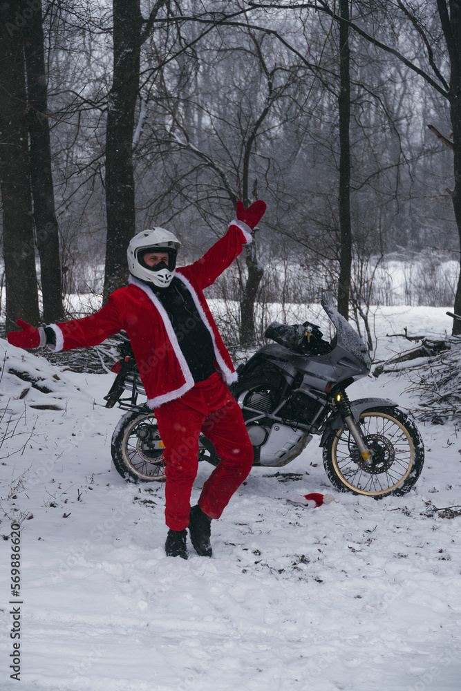 Happy motorcyclist in a Santa Claus suit shows likes. Winter forest with falling snow. Touring motorcycle in the background. The concept of New Year's holidays. Vertical photo.