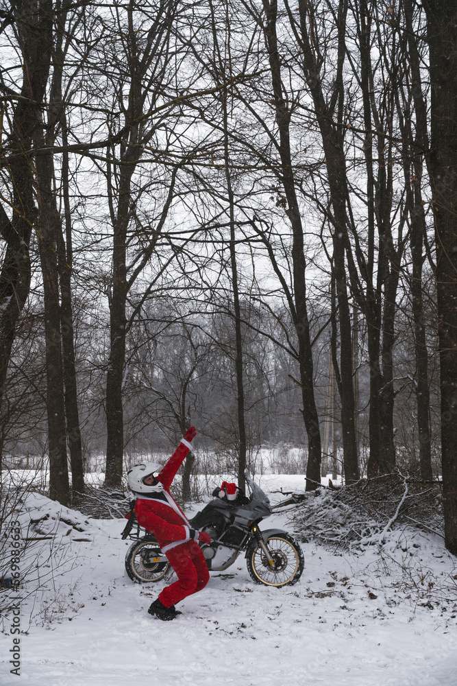 Happy motorcyclist in a Santa Claus suit shows likes. Winter forest with falling snow. Touring motorcycle in the background. The concept of New Year's holidays. Vertical photo.