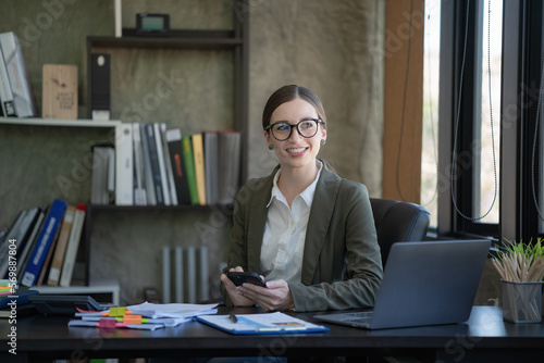 Businesswoman sits at a table in front of a laptop computer in her office and holds a phone in her hands and smiles. © Songsak C