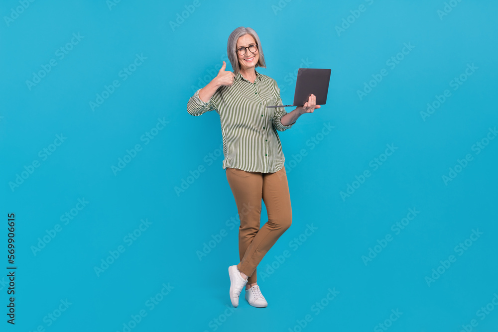 Full length photo of promoter programmer school education courses mature aged pensioner woman thumb up hold laptop isolated on blue color background Full length photo of promoter programmer school education courses mature aged pensioner woman thumb up hold laptop isolated on blue color background