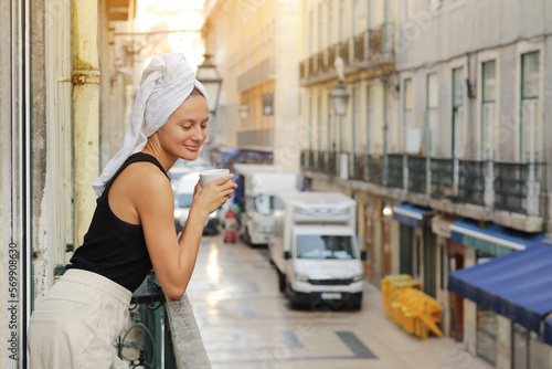 Young beautiful woman in a towel on head is drinking coffee or tea and smiling cute standing on balcony on ueropean street background. lifestyle morning. Concept of domestic or traveling lifestyle.