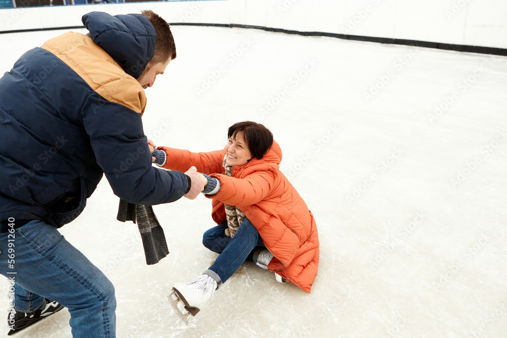 Middle-aged positive couple skating on open air ice rink. Man helping fallen woman to get up. Concept of leisure activity, winter hobby and sport, vacation, fun, relationship, emotions. Middle-aged positive couple skating on open air ice rink. Man helping fallen woman to get up. Concept of leisure activity, winter hobby and sport, vacation, fun, relationship, emotions.