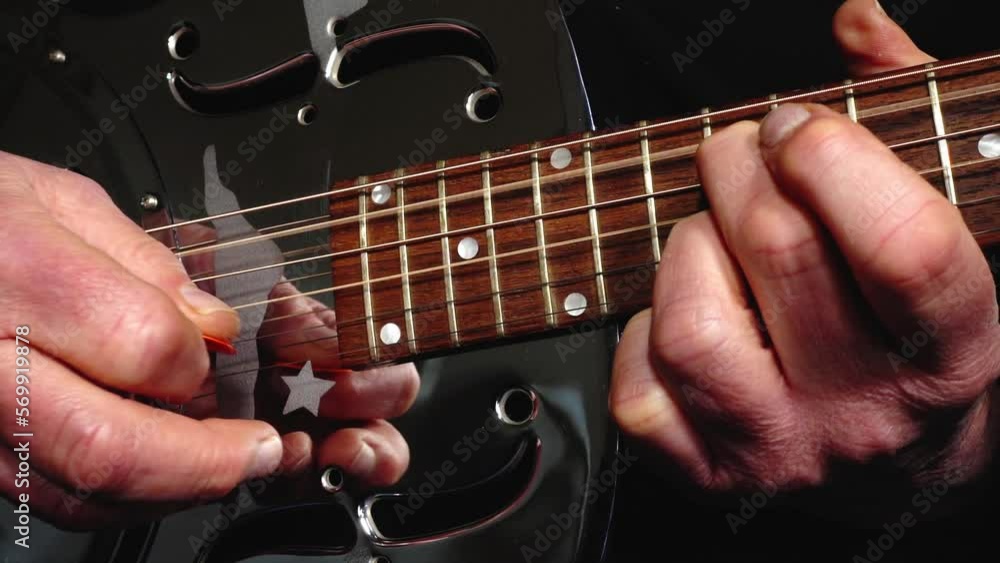Closeup of a man’s hands playing a shiny, metal body, resonator acoustic guitar, picking strings