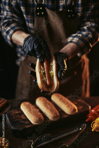 A guy in a leather apron makes a hot dog. The chef in black gloves puts a sausage in a bun.