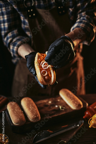 A guy in a leather apron makes a hot dog. A chef in black gloves puts an onion in a bun.