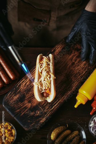 Hot dog cooking. The chef melts the cheese with a gas burner. Hot dog with Korean carrots and cabbage on a wooden board.