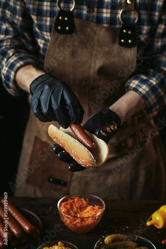 A guy in a leather apron makes a hot dog. The chef in black gloves puts a sausage in a bun.
