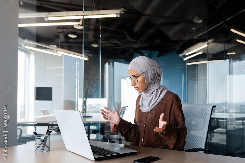 A young Arab woman in a hijab is sitting at a desk in the office and ...