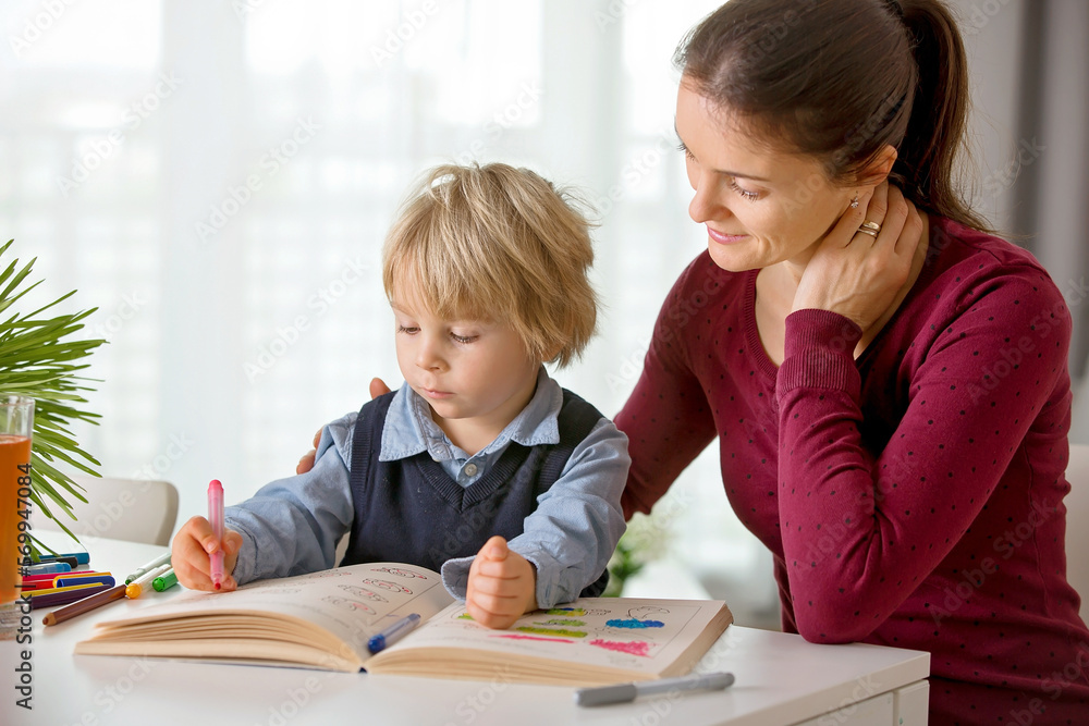 Cute preschool child, blond boy, filling some homework in a work book and coloring, mother helping him Cute preschool child, blond boy, filling some homework in a work book and coloring, mother helping him